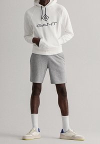 Sudadera blanca con el logo de GANT, pantalones cortos grises, calcetines blancos y zapatillas ligeras. La sudadera cuenta con cordones y mangas raglán.