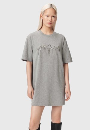 Ung kvinde med langt blondt hår iført en løs grå t-shirt-kjole med glitrende, dryppende kursiv tekst på fronten.