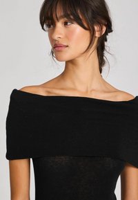 Juodas off-the-shoulder viršus, pagamintas iš minkštos medžiagos, prigludęs dizainas ir lygus paviršius, su plačiu iškirpimu ir minimaliu dekoru.
