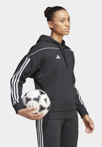 Zwart sweatshirt met witte strepen, capuchon en buidelzak, in combinatie met zwarte broeken, vasthoudend een zwart-witte voetbal.