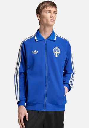 Giovane uomo che indossa una giacca con cerniera blu con strisce bianche sulle maniche e stemma della Svezia, in piedi con una mano nella tasca.