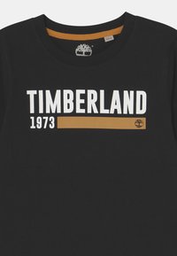 Svart bomullströja med en vit "TIMBERLAND" logotyp, "1973" i röd och ett guld horisontellt streck under texten. Rund halsringning.