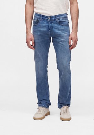 Homme portant un jean bleu droit, des baskets blanches et une chemise blanche à manches courtes, debout devant un fond uni.