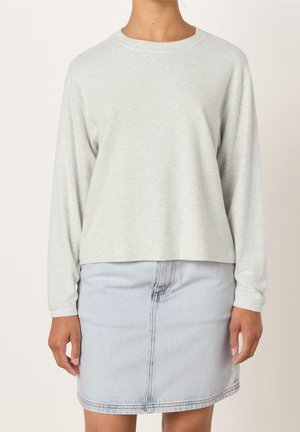 Personne portant une chemise à manches longues gris clair et une jupe en denim bleu clair, debout devant un fond uni.