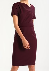 Gravidklänning i djup burgunder, med korta ärmar, en figurnära silhuett och en midjeaccent. Smoj g fabric med en lätt glans.