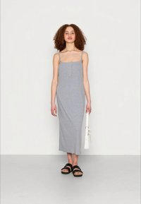 Mulher com cabelo encaracolado, vestindo um vestido midi cinza sem mangas e sandálias pretas, segurando uma bolsa branca ao seu lado.