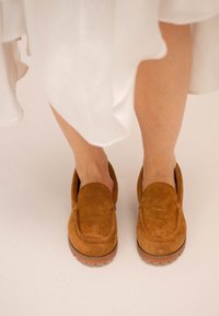 Brązowe zamszowe loafersy z okrągłym noskiem i grubą gumową podeszwą, częściowo widoczne spod zwiewnej białej odzieży.