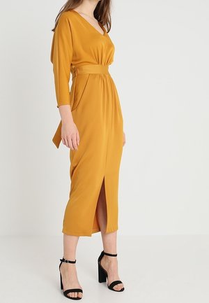 Vrouw draagt een mosterdgele midi-jurk met driekwartmouwen, V-hals, voorste split en zwarte sandalen met hoge hakken en enkelriem.