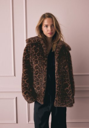 Jonge vrouw met lichtbruin haar draagt een bruine faux fur jas met luipaardprint over een zwart outfit, staand binnen tegen een lichte gepleisterde wand.
