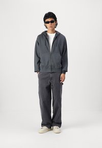 Dunkelgrauer Zip-Hoodie mit einer Fronttasche, kombiniert mit lockeren dunkelgrauen Cargo-Hosen und weißen Schuhen. Das Modell trägt schwarze Sonnenbrillen.