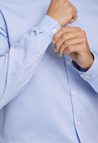 Camicia azzurro chiaro a maniche lunghe con una texture liscia, chiusura a bottoni sul davanti e bottoni bianchi a contrasto sui polsini e sul davanti.