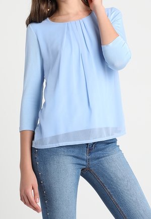 Femme portant un chemisier plissé bleu clair à manches 3/4 et un jean bleu orné de petits clous sur les côtés, posant avec la main près du visage.