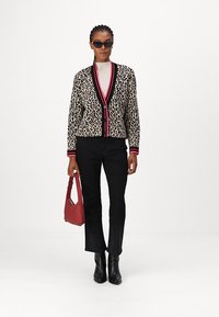 Karierte Leoprint-Cardigan mit pinken und schwarzen Akzenten, kombiniert mit schwarzen Hosen mit feiner Spitzenkante. Das Model hält eine rote Handtasche.