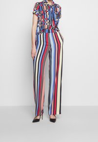 Chemisier coloré à motif avec des manches courtes et un nœud au cou, associé à un pantalon évasé à rayures de différentes couleurs. Talons noirs.