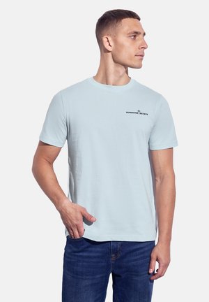 T-shirt en coton bleu clair à manches courtes, col rond, avec un logo noir sur la poitrine gauche. Porté avec un jean foncé.