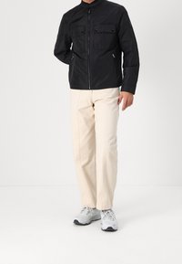 Černá bunda na zip s dvěma kapsami na hrudi a bočními zipy, doplněná světlými širokými kalhotami a šedými teniskami.