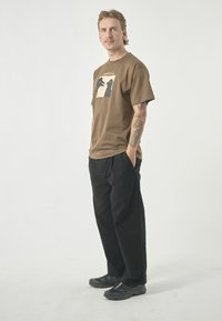 Brown cotton t-shirt featuring a graphic print on the front, paired with loose-fitting black pants. Rubber-soled black sneakers complete the look.