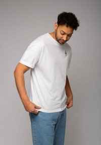 Weißes T-Shirt mit kurzen Ärmeln aus Baumwolle, versehen mit einem kleinen grünen grafischen Detail auf der linken Brust. Kombiniert mit hellblauen Jeans.