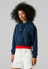 Mladá žena s kučeravými vlasmi, oblečená v námornícky modrej skrátenej mikine s nápisom "Red Bull Racing", pod ňou červený top a bielymi rifľovými šortkami.