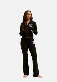 Zwarte zip-up hoodie en bijpassende broek, met witte en roze logo-details. De stof lijkt zacht en rekbaar, met een aansluitend ontwerp.