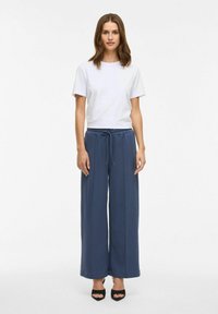 Witte T-shirt met korte mouwen, gecombineerd met donkerblauwe, wijde broek met een trekkoord in de taille, en zwarte open teen hakken op een neutrale achtergrond.