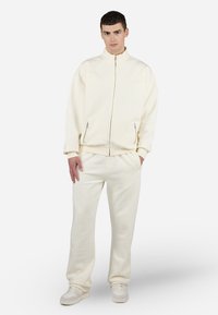 Hanorac crem cu fermoar și pantaloni de trening asortați, confecționați dintr-un material moale. Dispune de buzunare cu fermoar, manșete ribbate și broderie discretă cu logo.