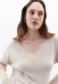 Beige gestrickter Pullover mit V-Ausschnitt, der über gerippte Akzente an den Ärmeln und einen leichten, strukturierten Stoff verfügt.