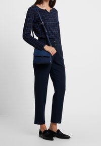 Granatowa bluzka w kratkę z długim rękawem, zestawiona z spodniami na wysoki talii w tym samym kolorze. Model trzyma małą, teksturowaną granatową torbę typu crossbody. Czarne loafersy.
