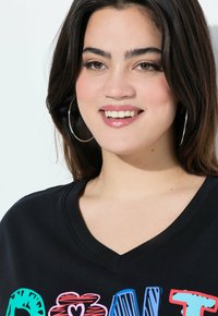 T-shirt nero a maniche corte con scollo a V e design grafico testuale colorato. Il modello ha i capelli lunghi e indossa grandi orecchini a cerchio.