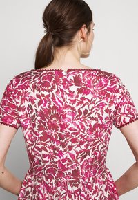 Femme aux cheveux bruns en queue de cheval, portant une robe à motif floral rose et rouge avec des bords festonnés, vue de dos.