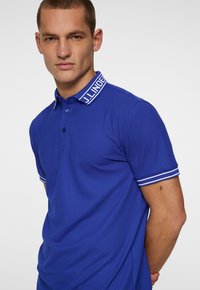 Blue polo shirt featuring a ribbed collar with “J. Lindeberg” logo, two buttons, and white striped accents on cuffs. Smooth fabric texture.