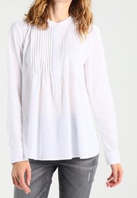 Blouse blanche à manches longues avec un devant plissé, un motif discret à pois texturé et un col mandarin avec une fermeture par bouton.
