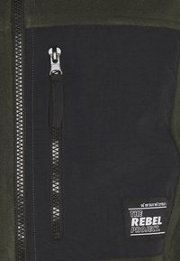 Fleece jacket section in dark green and black, featuring a zippered pocket, contrasting textures, and a branded label with white text.