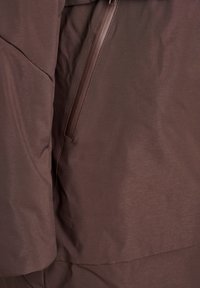 Gros plan sur un tissu de veste marron avec une poche zippée verticale et des coutures visibles sur la manche et le corps.