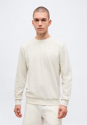 Młody mężczyzna z krótkimi włosami, ubrany w jasnobeżową bluzę i pasujące spodnie, stoi na jednolitym białym tle.