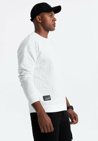 Vit sweatshirt med upphöjd textur och logotyp, rund halsringning, ribbade ärmslut och nederkant, matchad med svarta byxor och en svart keps.