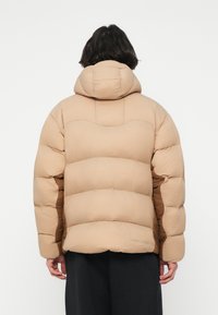 Chaqueta acolchada beige con un diseño de acolchado, capucha ajustada y mangas largas. Presenta una textura suave y un detalle del logo sutil cerca del dobladillo.