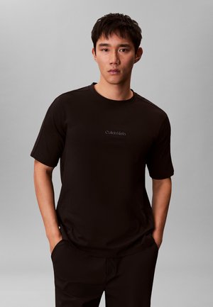 Jonge man die een zwart Calvin Klein T-shirt en zwarte broek draagt, met handen in de zakken, tegen een grijze achtergrond.