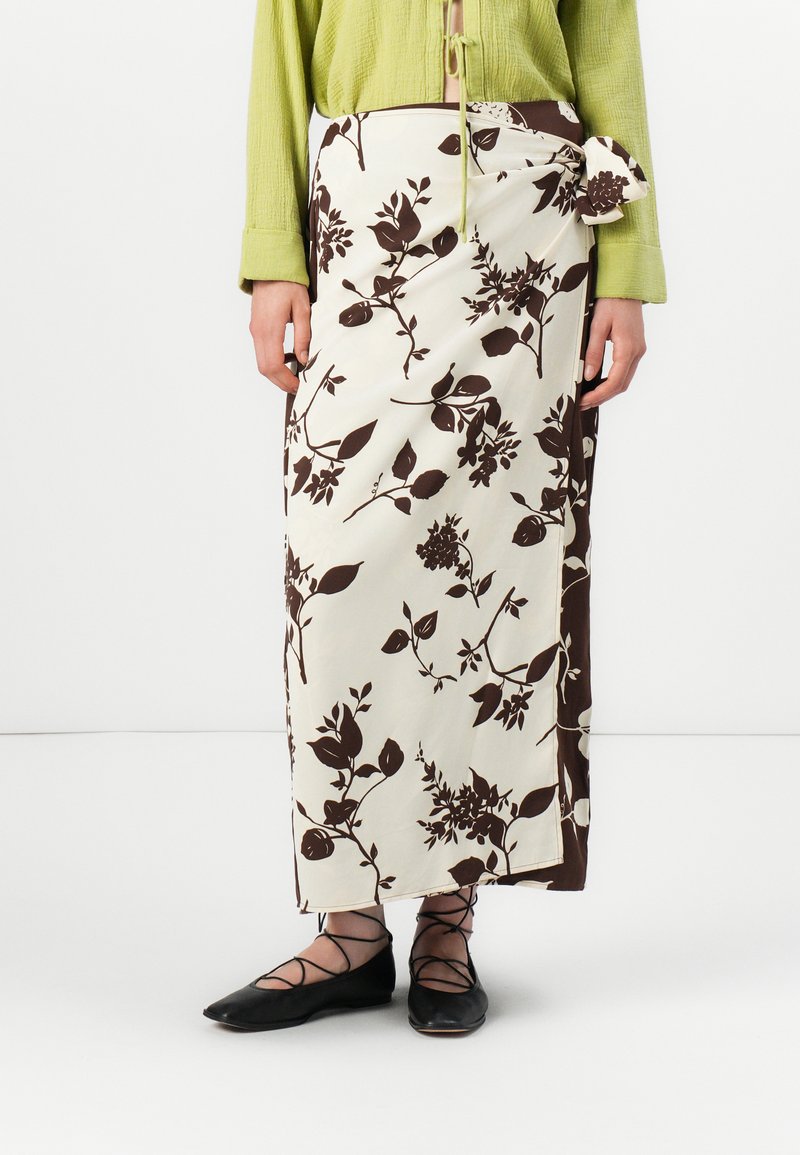 Jupe longue crème avec motif floral brun ; détail de noeud sur le côté ; associée à des chaussures plates noires avec des brides entrecroisées. La texture semble lisse.