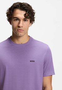 Violetti puuvillainen t-paita, jossa on lyhyet hihat, pyöreä pääntie ja musta "BOSS"-logo rinnassa. Sileä pinta, rento istuvuus.