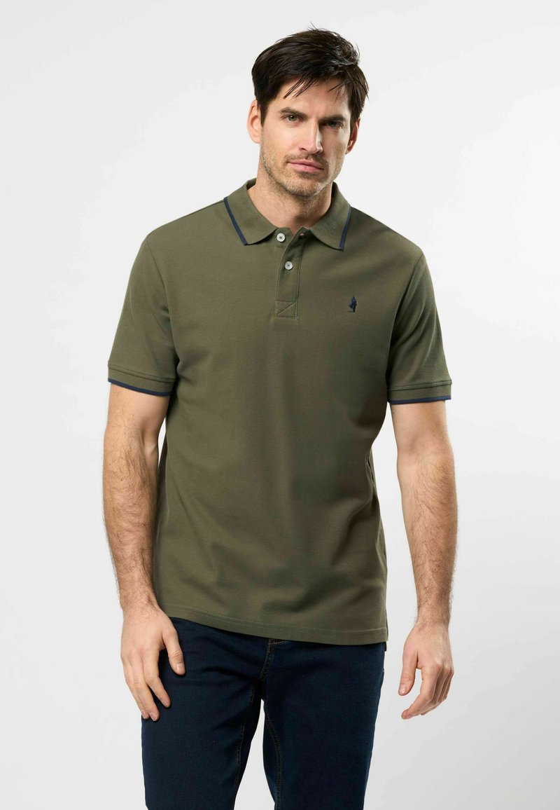 Homme debout devant un fond uni, portant un polo vert olive avec des détails bleu marine et un jean bleu foncé, faisant face à l'objectif avec une expression neutre.