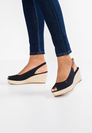 Pieds portant des sandales compensées bleu marine à bout ouvert avec des talons plateforme tissés et des brides cheville, associées à un jean denim bleu foncé.