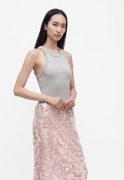 Jeune femme aux longs cheveux foncés, portant un haut gris sans manches et une jupe rose brillante à sequins, posant devant un fond blanc.