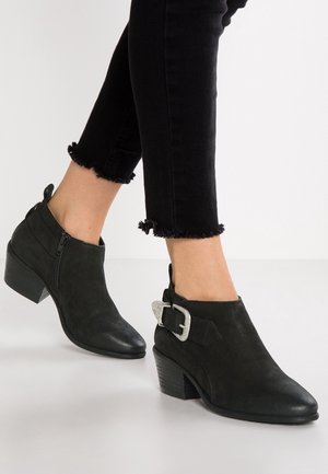 Bottines noires jusqu'à la cheville avec talons empilés bas, fermeture éclair latérale et détail de boucle argentée, portées avec un jean noir raccourci à ourlet effiloché.