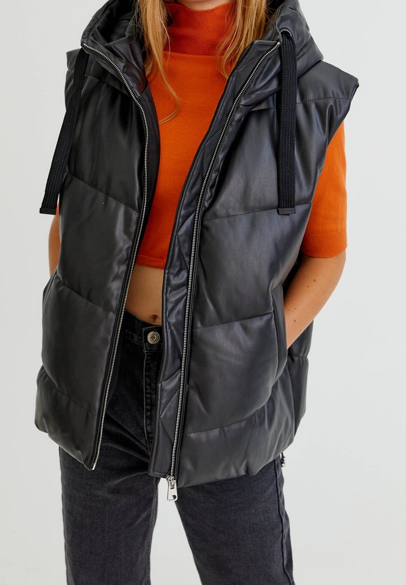 Veste matelassée noire avec capuche, en matériau synthétique brillant, fermeture éclair à l'avant et poches latérales, portée sur un haut orange.