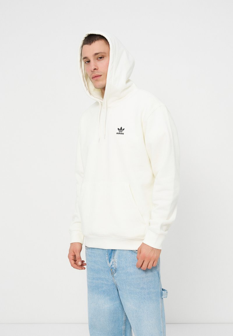 Mężczyzna ubrany w biały kapturowy sweter Adidas z założonym kapturem i jasnoniebieskie dżinsy, stojący na jednolitym białym tle.