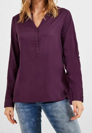 Woman wearing a long-sleeve purple blouse with a three-button V-neck paired with blue denim jeans against a plain background.