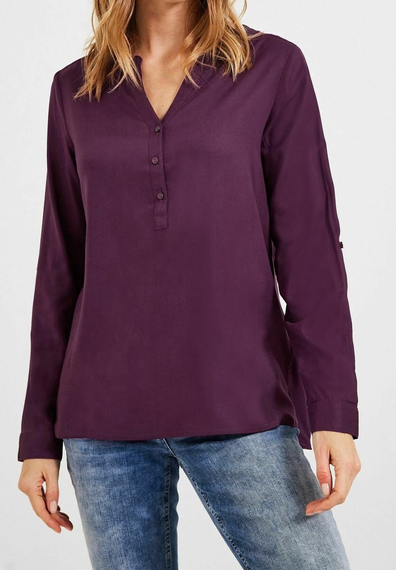 Femme portant un chemisier violet à manches longues avec un col en V à trois boutons, associé à un jean en denim bleu sur un fond uni.