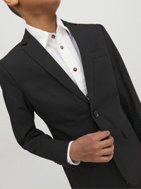 Zwarte blazer met enkele knoop, van bijpassend zwart materiaal en een sleek ontwerp. Witte blouse met kraag eronder met zichtbare bruine knopen.