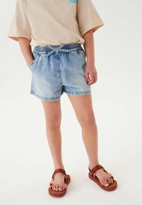Ljust blå denim shorts med elastisk midja, sidofickor och kontrasterande sömmar. Bärs med bruna och rosa sandaler.
